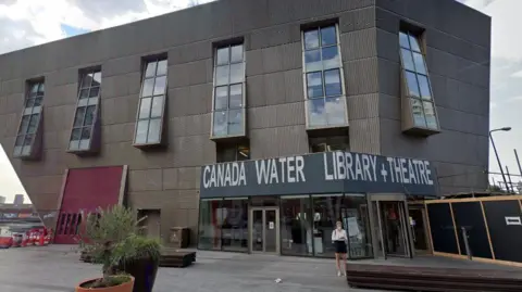 Google Inverted pyramid building with 'Canada Water Library and Theatre' sign on the right hand side. A woman stands in the foreground.