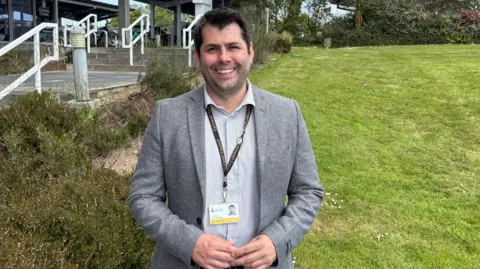 BBC Leigh Frost is smiling as he stands in a garden with County Hall behind him. He has dark hair and brown eyes and is wearing a grey jacket and a grey and white striped shirt and a lanyard with his Cornwall Council identification on it.