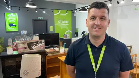 A man in a navy polo shirt and a lime green lanyard stands in a charity shop. The walls are white and dark grey with a large green sign reading 'Thank you Stone!' He is surrounded by used furniture such as cabinets, a lamp, chairs, television and keyboard