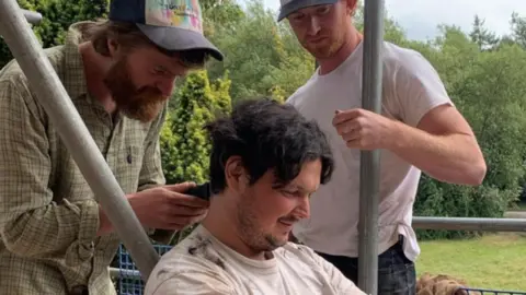 Tom Jones A man in jeans and t-shirt is sat on a workbench against some scaffolding.  He has medium length hair and a man behind him, wearing a shirt cap and jeans, us holding a shaver.  Another man in white tshirt and cap is watching.