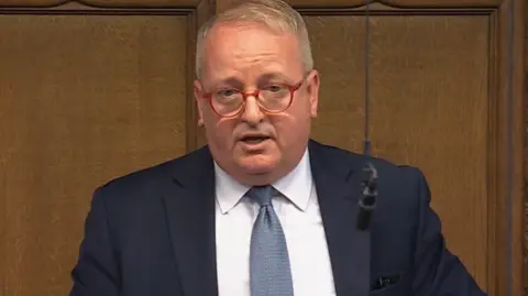 A man with short fair hair and red-framed glasses stands in the House of Commons in a navy blue suit, white shirt and blue tie. The wood panelling of the chamber is behind him, and a microphone hangs on a wire in front of him.
