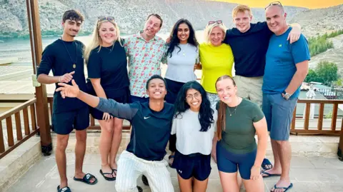 BBC/Studio Lambert Kush, Katie, Mark, Jo, Puja, Roshni, Margo, Molly, Harrison, Andrew pose as a group on a balcony in Halfeti, Turkey with a river and mountain range behind.