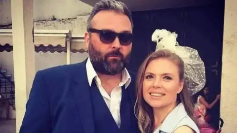 Jamieson Hill A couple stand with their arm around one another. Hill is wearing a blue suit and sunglasses. He has short grey hair that is swept backwards and a black beard. Şeyda has long wavy brown hair and has a white hat and light blue dress on.