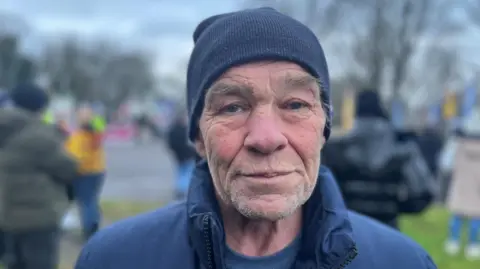 Mr Glass is an older man and he is wearing a blue hat and a blue jacket.