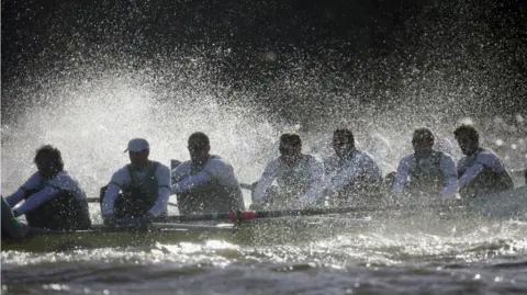 Getty Images Seven male rowers can be seen in white bibs and sunglasses in a yellow scull. There is significant splash from the river being sprayed around the rowers, and the sunlight is reflecting off the water.