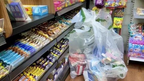 Three bags of vapes next to a counter with lots of chocolate bars and other sweets on the counter of the shop. They are all different colours.