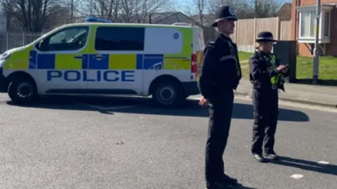 Two police officers stand in a road. They are wearing black police uniform and hats. There is a police van with yellow and blue on the side, and the word POLICE written on the side.