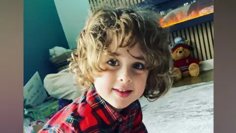 Contributed A young boy with brown curly hair smiles at the camera as he plays on the floor of a living room. He wears a chequered red shirt. 