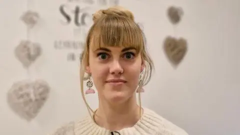 BBC / Mairead A young woman with blonde, with a full fringe and her hair tied up, wearing decorative earrings and a cream jumper.