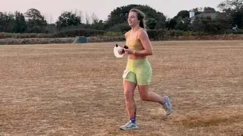 Emily Torode Emily Torode wears a yellow crop top and green leggings as she runs across a field 