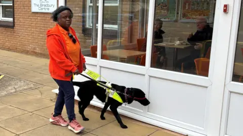 Naomi has medium length black and grey hair and is wearing a hairband, orange t-shirt, red jacket, blue trousers and pink trainers. She is walking outside The Branbury Centre with a black guide dog which has a yellow harness.