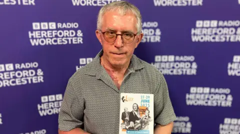 A man stands in front of purple board holding a leaflet.
