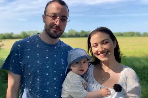 Kianty and Conor smile at the camera. They hold Luke, who wears a blue sunhat. There is a yellow field and a hedgerow in the background. 