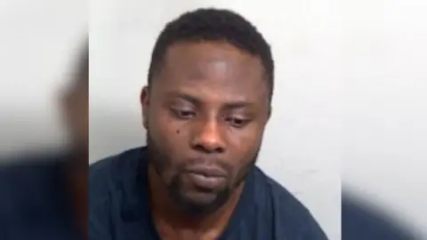 Will Dohmatob is in the middle of the image. He has black short hair and is wearing a navy T-shirt. He is looking toward the floor to the right and sat in front of a white background.