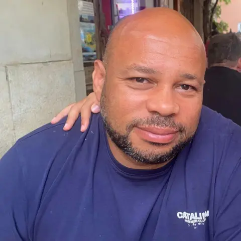 Matthew Lasebikan Matthew, in a purple t-shirt smiles at a camera. He is bald and has short stubble. It is a head and shoulders shot.