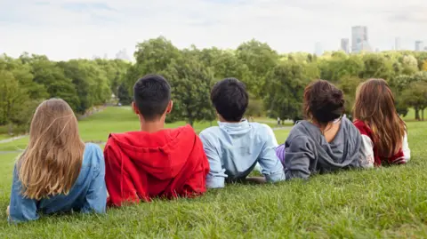 Five boys and girls lie on a hill looking down over parkland. Their backs are to the camera. They have hooded tops and/or casual clothing on. Trees line part of the park with tower blocks visible in the background on the right-hand side. 