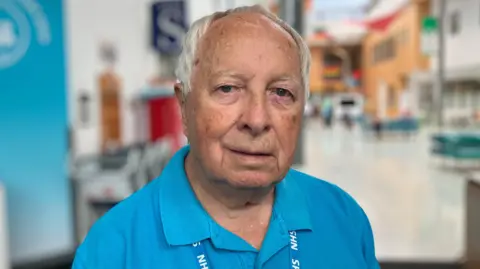 Shariqua Ahmed/BBC Ronald Nash has white hair and is wearing a blue T-shirt and NHS lanyard. He is looking into the camera. The background behind him is blurred.