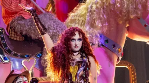 Getty Images Chappell Roan performing on stage during The Grammy's. She wears a metallic pink and yellow body suit with matching elbow-length gloves. She holds her right hand up as she looks at the crowd, standing in front of an enormous pink horse model with a blonde mane. 