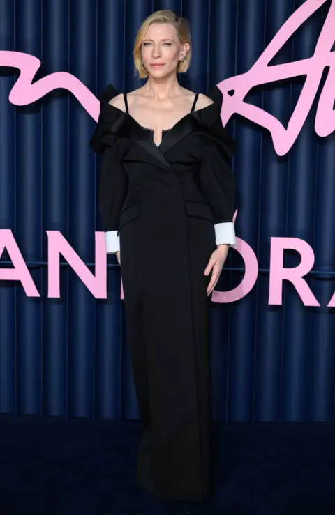 Getty Images A full-length view of Cate Blanchett in black dress with white sleeves, hand clasped together, standing in front of a blue curtain background
