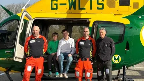 Family photo People in front of a colourful air ambulance helicopter, which is painted green and yellow. The helicopters side doors are open with two teenage boys sitting smiling at the camera. Standing beside them are three men - two in uniforms of orange and black and other dressed in black fleece and trousers.