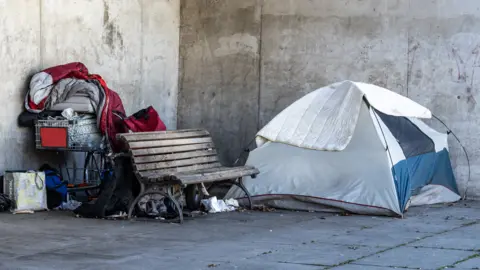 A tent and a supermarket trolley loaded with posessions are either side of a bench on an urban street.