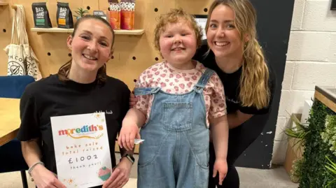 Family handout Two women stood smiling with one holding a piece of paper that says Meredith's Mission bake sale total raised £1002 thank you!! and a girl in the middle wearing blue dungarees.
