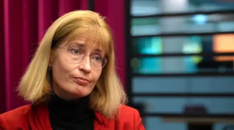 A woman with shoulder-length blonde hair, frameless glasses, a red jacket and black shirt looks slightly to her left, tilting her head
