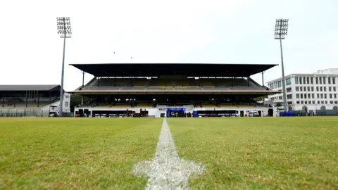 Getty Images A view of a two-tiered sport stadium stand. In the foreground is a green sports pitch marked with white paint to indicate its centre spot.