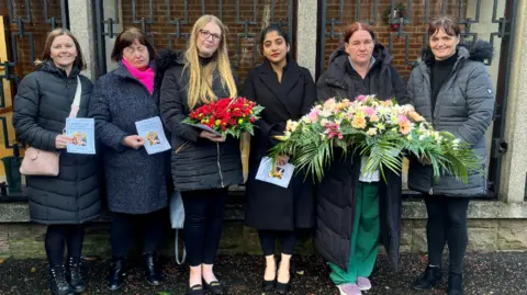 Six ladies pose for a photo holding funeral flowers, and the orders of service from the funeral of Ann Hillman. They are all wearing black or grey raincoats. 