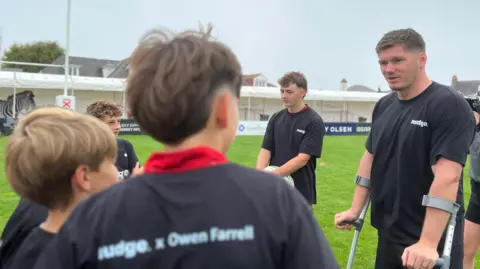 BBC A group of young rugby participants wearing black Nudge-branded shirts stand in a circle on a rugby pitch during a coaching session. Owen Farrell is using forearm crutches while he addresses the group. Rugby club buildings and goalposts are visible in the background under an overcast sky.