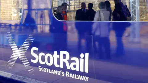 A ScotRail train sitting in a station. A group of people talking can be seen reflected in a window.