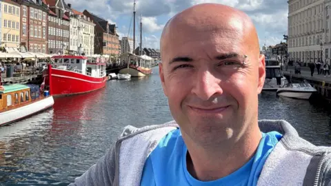Gray Davis Gray Davis smiling slightly at the camera in front of a dock with boats parked in the water on a sunny day