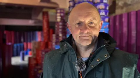 Teddy's grandfather Tim smiling for a picture as he is surrounded by towers of tubs behind him.