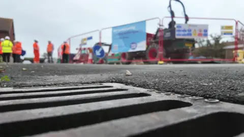 A drain in the ground with people in high visibility clothing in the distance working next to fencing and construction machinery. 