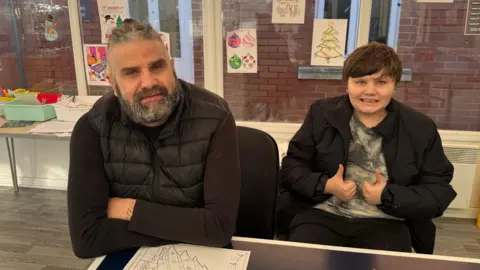A man with a long salt and pepper beard, and similar-coloured hair, wearing a black body warmer and sitting at a table. A young boy is sitting next to him with his thumbs up to the camera.