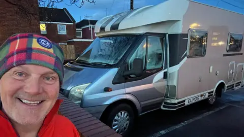 David Matthews A man smiles for a selfie in-front of a large silver camper van. He is wearing a fleece head scarf