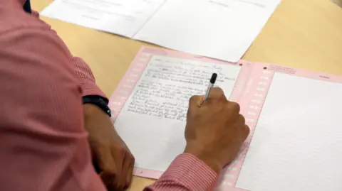 An anonymous hand writes on notepaper against a wooden desk.