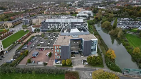 Steve Hubbard/BBC Aerial shot of the UON Waterside Campus, showing river to the right, and modern grey and white university buildings to the left. There are houses in the background.