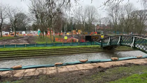 Nottingham City Council View across a canal/river towards a grassy area which is undergoing substantial ground work, with barriers, fences and machinery visible.