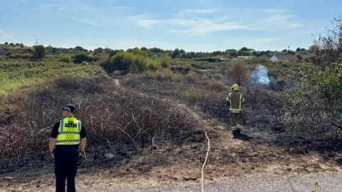 DWFRS A firefighter and a police officer stand in front of a smouldering area of vegetation