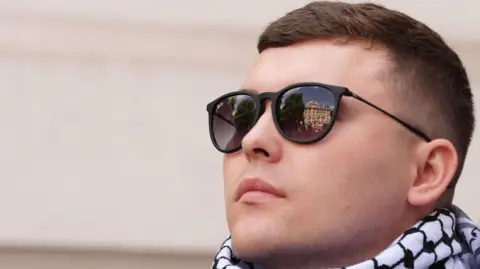 A close-up of a man in the midst of a crowd. He is wearing sunglasses and a black and white keffiyeh headdress around his neck like a scarf. There is a camera lens being held close to his face as a photographer takes a photo