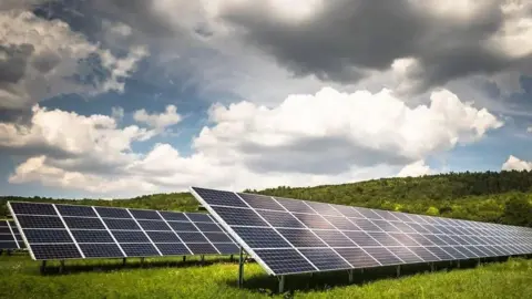 Two large ground-mounted solar panels are pictured in a green field facing towards the sky which is a mix of cloud and sun.

