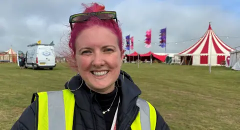 Llio is standing at the festival site; behind her is a red and white striped tent, festoon lights and pink and purple flags. She has bright pink hair and green sunglasses on top of her head. She has large silver hoops in her ears and is wearing a silver necklace featuring stars, a yellow high-vis vest and a blue coat.