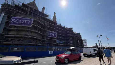The Old College surrounded by scaffolding, two people walk by looking at the works.