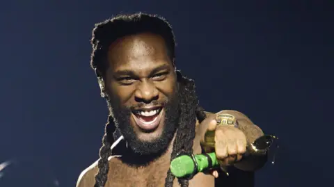 Burna Boy pointing with his finger, holding a green microphone