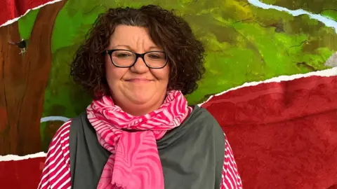 Julie Graham - a woman with curly brown, shoulder length hair, wearing glasses a grey top with a pink scarf, pictured in front of a mural showing a tree