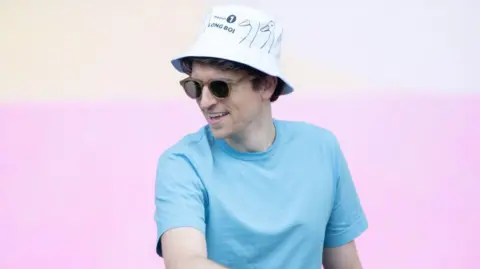 Getty Images Greg James performs a DJ set in front of a pastel pink backdrop on-stage at Big Weekend. He wears a light blue t-shirt, sunglasses, and a white bucket hat printed with line-drawn pictures of a duck, the Radio 1 logo and the words "Long Boi" in black letters.