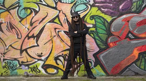 Borkowski Arts Alborosie press picture of the artist looking towards the camera in a posed stance with a graffiti background 