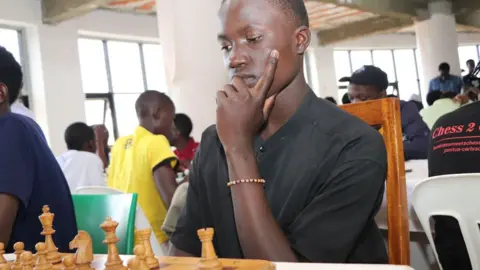 Wycliffe Muia / BBC Jovan Kasozi in a black jersey top and wearing a bracelet stares in concentration, his hand on his chin, at a chess board with brown wooden pieces in a room where a chess tournament is being held.