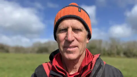 A man is part smiling at the camera. He has a orange and black bobble hat and is wearing a red t-shirt and fleece and a National Trust jacket. 